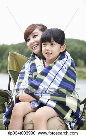 Stock Image - a mother and a daughter sitting on mother's lap having a blanket together. Fotosearch