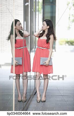 A woman in coral dress and grey clutch looking herself through a glass wall View Large Photo Image Picture - A woman in coral dress and grey clutch looking herself through a glass wall. Fotosearch