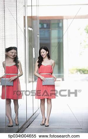 Stock Photography - A woman in coral dress and grey clutch walking along a glass wall. Fotosearch