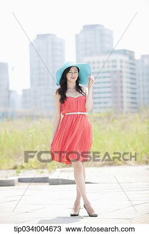 Stock Image - A woman in coral dress and turquoise hat posing in front of grass field. Fotosearch