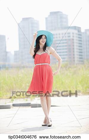 Stock Photograph - A woman in coral dress and turquoise hat posing in front of grass field. Fotosearch