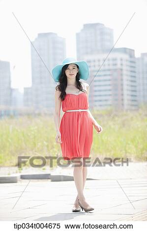 Stock Photography - A woman in coral dress and turquoise hat posing in front of grass field. Fotosearch