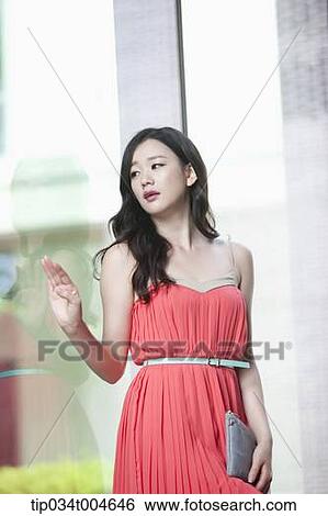 Stock Photograph - A woman in coral dress posing next to a glass wall. Fotosearch