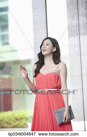 Stock Photo - A woman in coral dress smiling next to a glass wall. Fotosearch