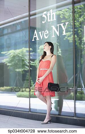 Stock Photo - a woman in coral dress standing in front of a show window with a 5th Ave NY sign. Fotosearch