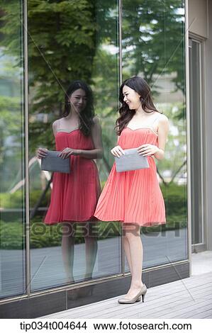 Picture - A woman in coral dress walking next to a glass wall. Fotosearch