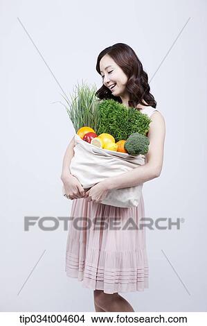 Picture - A woman in dress holding a bag of vegetables. Fotosearch