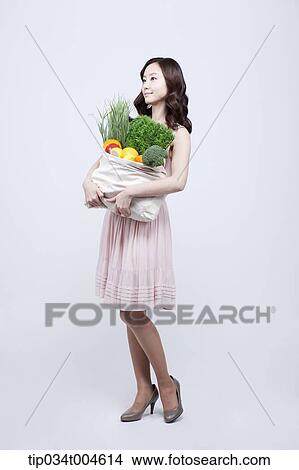 Picture - A woman in dress holding a bag of vegetables. Fotosearch