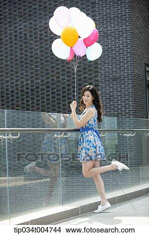 Picture - a woman in flower printed outfit posing with a bunch of balloons in a building balcony. Fotosearch