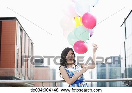 Stock Photo - a woman in flower printed outfit posing with a bunch of balloons in a building balcony. Fotosearch