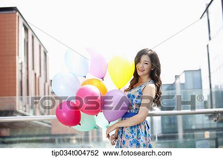 Stock Image - a woman in flower printed outfit hugging a bunch of balloons in a building balcony. Fotosearch