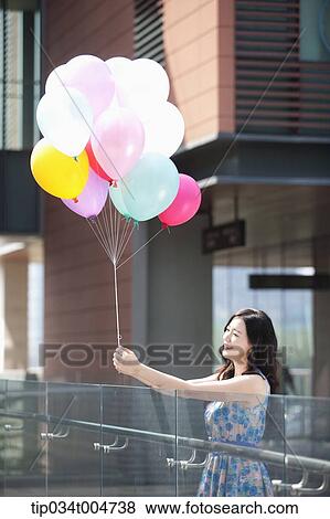 Stock Photo - a woman in flower printed outfit holding a bunch of balloons in a building balcony. Fotosearch