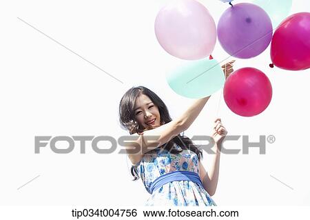 Stock Photograph - a woman in flower printed outfit holding a bunch of balloons in white background. Fotosearch