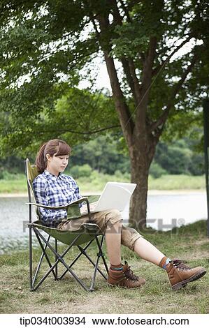Picture - a woman sitting on a camping chair using laptop. Fotosearch