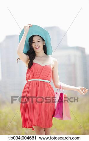 Stock Image - A woman smiling in coral dress and turquoise hat holding a shopping bags. Fotosearch
