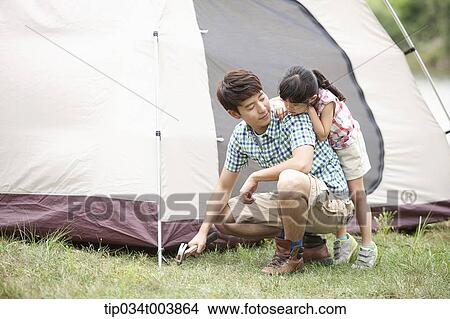 Picture - daughter leaning on back of her father hammering to build a tent. Fotosearch
