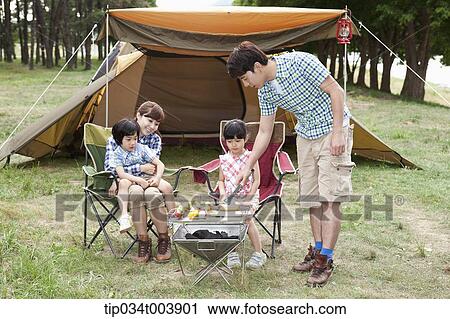 Stock Image - father grilling while other family members sat on camping chairs . Fotosearch