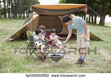 Stock Image - father grilling while other family members sat on camping chairs . Fotosearch