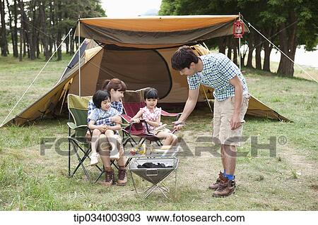 Stock Image - father grilling while other family members sat on camping chairs . Fotosearch