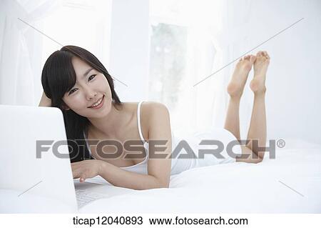 Stock Image - woman wearing white sleepwear and using the laptop on the bed . Fotosearch