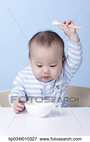 Stock Photo - a baby with a bowl and a spoon on table. Fotosearch