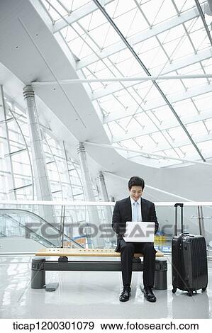 Stock Photo - a businessman using laptop while sitting on a bench in the airport. Fotosearch