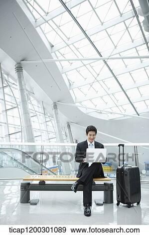 Stock Photo - a businessman using laptop while sitting on a bench in the airport. Fotosearch