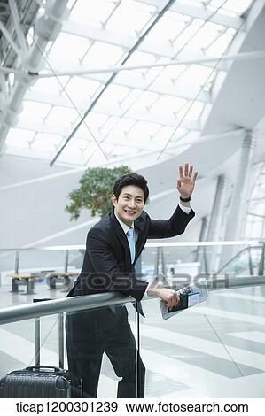Stock Photo - a businessman with suitcase greeting while leaning on fence in the airport. Fotosearch