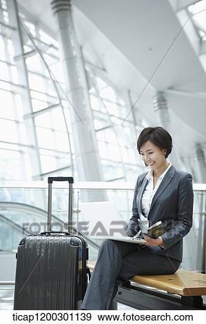 Stock Photo - a businesswoman using laptop while sitting on a bench in the airport. Fotosearch