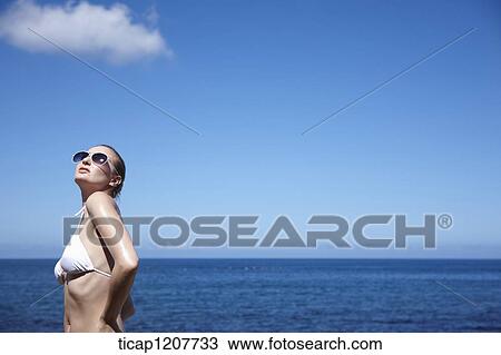 Stock Image - a lady in bikini and sunglasses posing under bright sky. Fotosearch