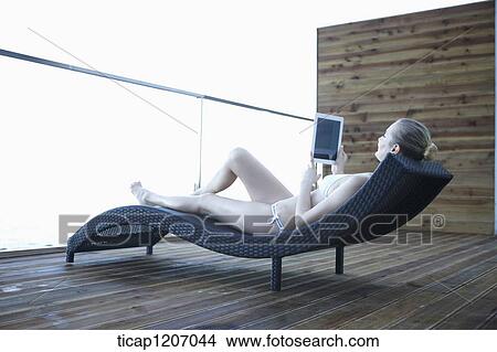 Picture - a lady in bikini lying on a beach bed using tablet PC. Fotosearch