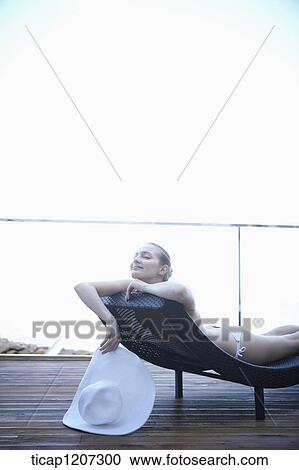 Stock Image - a lady in bikini lying on a beach bed holding beach hat. Fotosearch