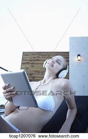 Stock Photograph - a lady in bikini lying on a beach bed listening to a music. Fotosearch