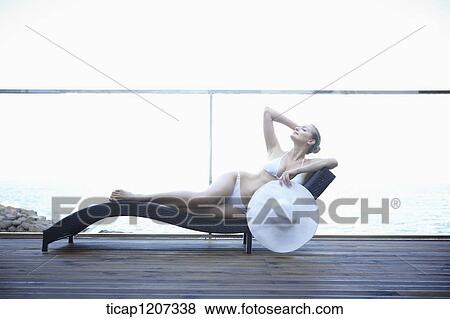 Stock Photo - a lady in bikini posing on a beach bed holding beach hat. Fotosearch