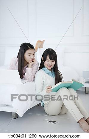 Stock Image - a lady using a laptop while other lady is reading a book. Fotosearch