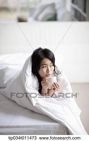 Stock Image - a woman drinking cup of coffee while wrapped in blanket. Fotosearch
