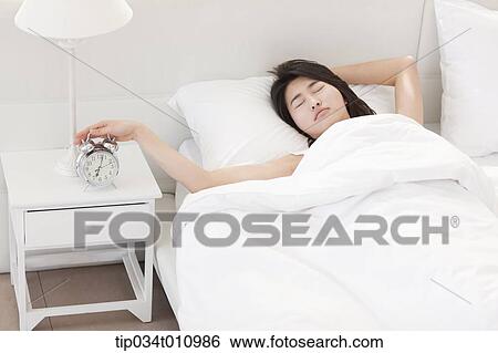 Stock Photograph - a woman on the bed with a hand on alarm clock. Fotosearch