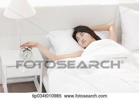 Stock Photography - a woman on the bed with a hand on alarm clock. Fotosearch
