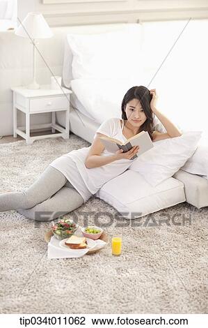 Stock Image - a woman reading a book next to a bed. Fotosearch