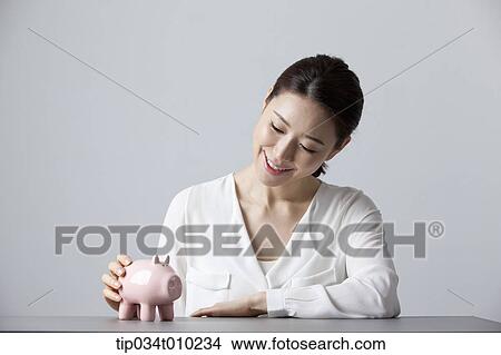 Picture - a woman smiling at desk with a piggy bank . Fotosearch
