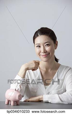 Stock Image - a woman smiling at desk with a piggy bank . Fotosearch