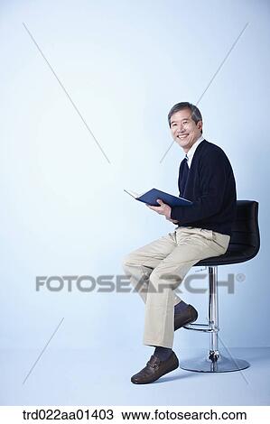 Stock Image - middle aged man reading a book while sitting on a chair. Fotosearch