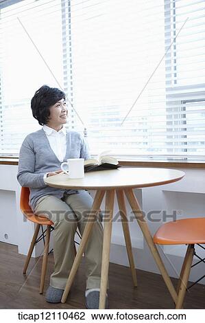 Stock Image - middle aged woman reading a book sitting at table. Fotosearch