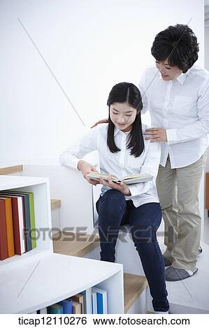 Stock Photograph - mother standing next to a student sitting on bookshelf reading. Fotosearch
