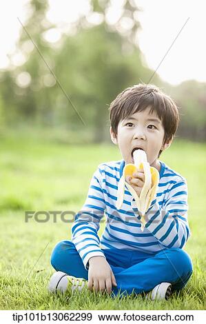 Stock Photo - a boy sitting on the grass eating a banana. Fotosearch