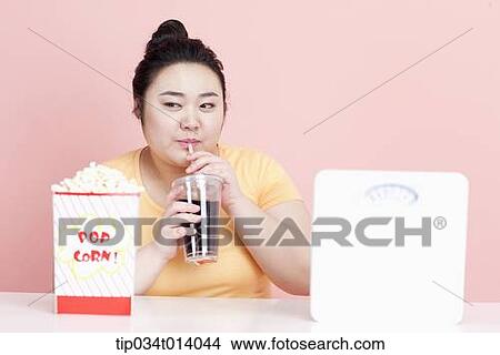 Picture - a chubby woman drinking a coke with popcorn in front of her. Fotosearch