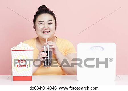 Stock Image - a chubby woman drinking a coke with popcorn in front of her. Fotosearch