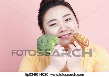 Stock Image - a chubby woman holding broccoli and cupcake. Fotosearch