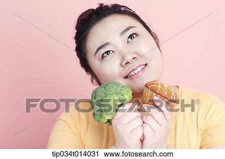 Stock Image - a chubby woman holding broccoli and cupcake. Fotosearch