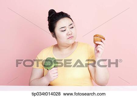 Stock Photo - a chubby woman holding broccoli and cupcake. Fotosearch